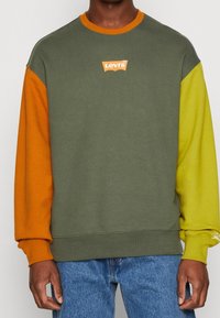 Felpa verde oliva con maniche arancioni e giallo brillante. Presenta un piccolo logo Levi's arancione sul davanti. Tessuto in misto cotone.