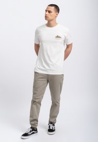 Witte korte mouwen t-shirt met een kleine graphic op de borst, gecombineerd met lichtgrijze broek en zwart-witte sneakers.