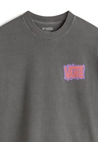 Camiseta gris de corte holgado con cuello redondo. Presenta un logo de Vans en morado con un estilo de papel rasgado en el lado izquierdo del pecho. Textura suave.