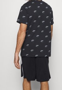 Černé tričko s opakujícími se bílými logy a textem, spárované s černými šortkami s bílými bočními pruhy. Bavlněný materiál, krátké rukávy.