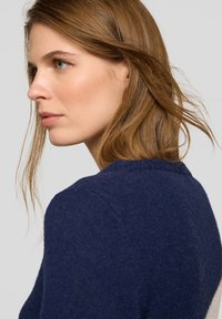 Marineblauer, gestrickter Pullover mit strukturierter Oberfläche, ausgestattet mit einem Rundhalsausschnitt und dezenten Schulternähten, getragen von einem Modell mit langen Haaren.