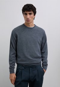 Grå stickad tröja med rund halsringning och långa ärmar, kombinerad med mörka byxor med veck fram och knappdetaljer.