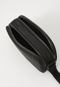 Sac en nylon noir à fermeture éclair avec trois compartiments, doté de fermetures éclairs en métal argenté et d'une sangle en tissu tissée pour le transport.