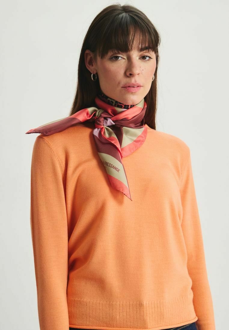 Pull en tricot orange avec un ourlet côtelé, associé à une écharpe en soie multicolore ornée de motifs roses, beiges et violets nouée autour du cou.