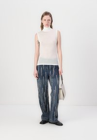 Ermeløs hvit genser med høy hals og gjennomsiktig finish, parret med blå vide jeans med vertikale sølvstriper.