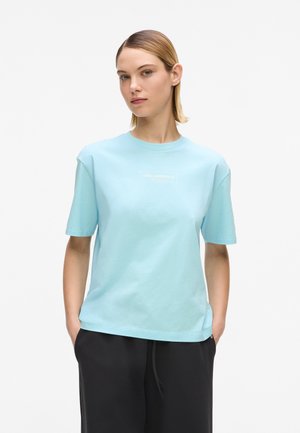 Femeie purtând un tricou albastru deschis cu mâneci scurte și un logo alb subtil pe piept și pantaloni negri, stând cu mâinile în buzunare.