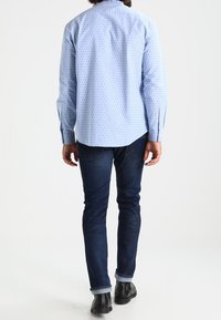 Camisa azul claro con pequeños puntos en azul oscuro, con cuello abotonado y mangas largas. Combinada con jeans azul oscuro y botas negras hasta el tobillo.