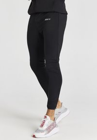 Černé sportovní jogger kalhoty s úzkým střihem, které mají decentní logo na stehně a hladkou, elastickou látku. V kombinaci se šedými a červenými teniskami.