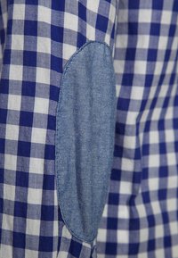 Niebiesko-biała tkanina w kratę w wzór gingham z łatek na łokciu z denimu. Łatka ma owalny kształt i gładką powierzchnię, kontrastującą z teksturowanym, kratkowanym materiałem.