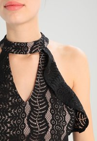 Haut en dentelle noire à col halter, avec un décolleté en trou de serrure et des motifs floraux, en tissu texturé et avec un détail de manches volantes.