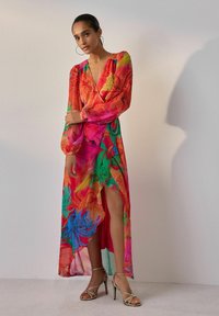 Rochie maxi floral colorată, cu decolteu adânc în V, mâneci lungi tip puff și fanta înaltă, realizată dintr-un material ușor. Sandale cu toc aurii.
