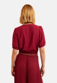 Bordeauxfarbene Bluse mit Puffärmeln, strukturiertem Stoff und rundem Ausschnitt. Kombiniert mit plissierten Hosen, die an der Taille mit einem goldakzentuierten Gürtel gerafft sind.