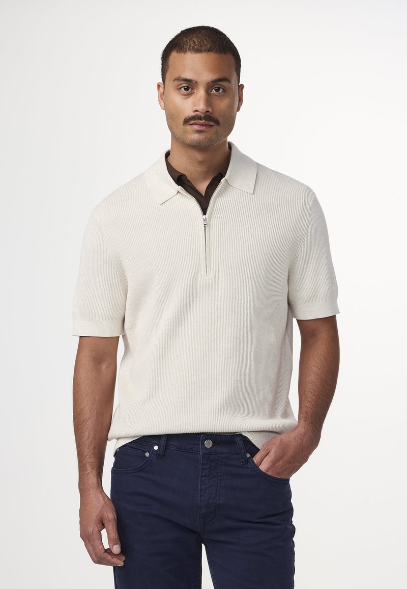 Sweter polo w kolorze kremowym z zamkiem błyskawicznym w kołnierzu i prążkowaną fakturą, w połączeniu z ciemnoniebieskimi dżinsami.