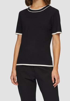 Kvinde iført sort kortærmet top med hvide kanter på krave, ærmekanter og kant, kombineret med sorte bukser mod grå baggrund.