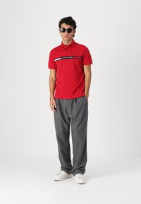 Rød polo skjorte med kontrasterende sorte og hvide striber, udstyret med 'TOMMY HILFIGER' logo. Grå bukser og hvide sneakers fuldender looket.