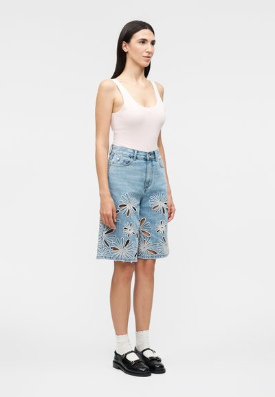 Γυναίκα που φοράει λευκό τοπ, τζιν σορτς με ψηλή μέση μέχρι το γόνατο με μεγάλα λουλουδένια κοψίματα, λευκές κάλτσες και μαύρα loafers.