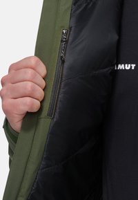 Giacca verde con interni neri isolati, dotata di una tasca con zip sul lato sinistro. Il tessuto è liscio, con dettagli di cucitura ben visibili.