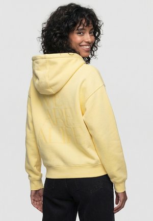 Woman with curly hair smiling over her shoulder, wearing a pale yellow hoodie with embossed text and black jeans.