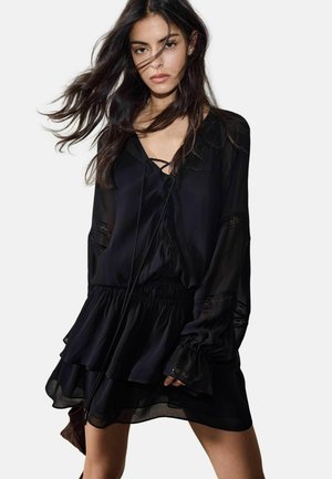 Frau in einem schwarzen, langärmeligen Kleid mit gerafftem Saum und Spitzenapplikationen, deren Haare ihr ins Gesicht wehen, vor einem schlichten Hintergrund stehend.