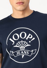 T-shirt en coton bleu marine avec un grand logo graphique blanc comportant le texte "JOOP!" et "JEANS", intégrant un design ressemblant à des éléments végétaux.