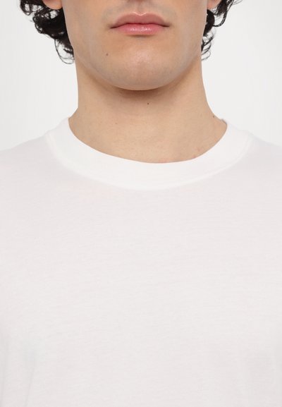 Tricou alb din bumbac, cu guler rotund, textură netedă și detalii de cusătură subtile de-a lungul gulerului și cusăturilor umerilor.