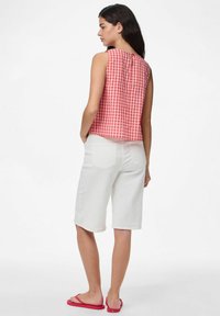 Červená a bílá kachlová bezrukávová blůza se rovnými liniemi a knoflíkovým detailem na zádech, v kombinaci s bílými šortkami ke kolenům a růžovými žabkami.