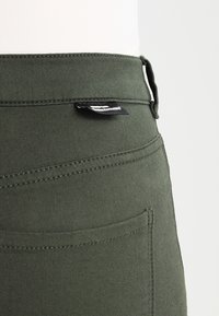 Pantalones verdes oliva hechos de una tela suave con una textura lisa, con cinturilla, bolsillo trasero y una pequeña etiqueta de marca adjunta.