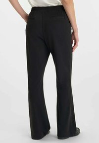 Persoon in een zwart, elastische taille, wijde broek, staand met één hand op de heup tegen een effen achtergrond.