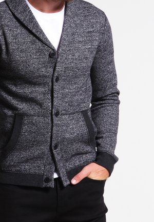 Mężczyzna w szarym kardiganie na guziki noszącym białą koszulę, z jedną ręką w kieszeni na białym tle.