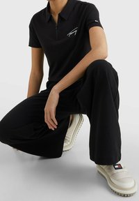 Černé polo tričko se zipem vpředu a bílým logem. Kombinováno s volnými černými kalhotami a krémovými botami s texturovanou podrážkou.