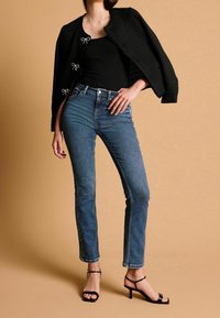 Veste noire texturée avec des accents de nœud, portée sur un haut noir ajusté. Jean bleu foncé et talons noirs à brides complètent la tenue.