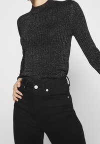 Czarna prążkowana bluzka z długim rękawem z metalicznymi nitkami, z wysokim kołnierzem, dopasowanym krojem. W zestawie z czarnymi dżinsami z wysokim stanem i przednimi kieszeniami.