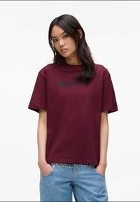 Jovem mulher com cabelo longo e escuro usa uma t-shirt oversized bordeaux com o texto "Karl Lagerfeld Jeans" e jeans de denim azul claro com cinto de tachas.