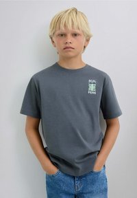Niño rubio con las manos en los bolsillos, vistiendo una camiseta gris con un gráfico en verde-azul y jeans azules, de pie contra una pared clara.