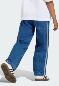 Jeans di denim blu con vestibilità rilassata, cuciture chiare e strisce laterali. Abbinati a scarpe bianche con strisce nere e suola marrone.