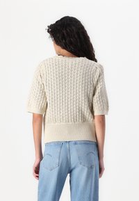 Knit sweater in cream with diamond texture, short sleeves, and ribbed hem. Paired with light blue high-waisted jeans, showing back view.