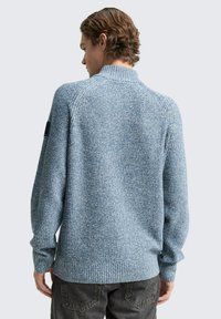 Lichtblauwe gebreide trui met textuur, voorzien van een hoge kraag, ribboorden en zoom, met een klein zwart embleem op de linkerschouder.