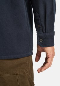 Marineblauwe langesleeve shirt met een gestructureerde afwerking, knoopmanchetten en contrasterende stiksels, gecombineerd met bruine corduroybroeken.