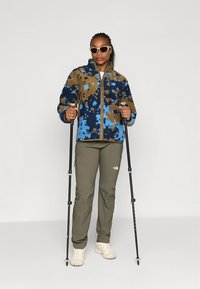 Giacca in pile con motivo blu e marrone con cerniera, abbinata a pantaloni verde oliva e calzature bianche. Include bastoncini da trekking e occhiali da sole.