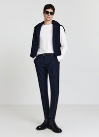 Marineblaue Wollweste über einem weißen Langarmhemd, kombiniert mit maßgeschneiderten dunklen Hosen und glänzenden schwarzen Stiefeletten. Accessoires umfassen schwarze Sonnenbrillen.