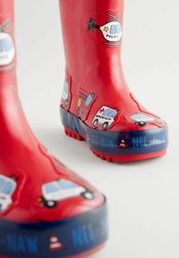 Bottes de pluie en caoutchouc rouge avec des motifs de véhicules d'urgence en cartoon ; présente une bande bleue à motif avec le texte "NEE-NAW". Semelle texturée pour une meilleure adhérence.