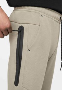 Beige Jogginghosen mit texturierter Oberfläche, ausgestattet mit einer eleganten schwarzen Reißverschlusstasche und Kordelzug, die funktionale Designelemente hervorhebt.