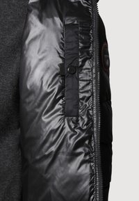 Sort polstret jakke med quiltet tekstur, frontlynlås, knaplukket lomme og et logo-patch på siden. Glat, letvægtsmateriale.