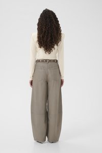 Kobieta o długich kręconych włosach, ubrana w kremową bluzkę z długim rękawem i spodnie w kolorze taupe o wysokim stanie i szerokich nogawkach z paskiem z ćwiekami, stojąca tyłem.