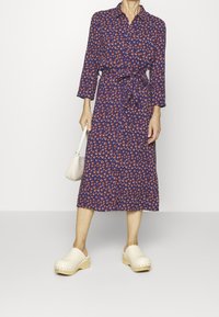 Abito midi con stampa floreale in navy, decorato con pois rossi e bianchi, vita con cintura, maniche lunghe e colletto tondo. Abbinato a zoccoli beige.