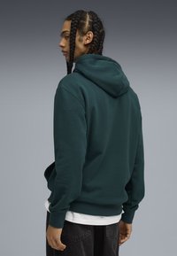 Sudadera verde oscuro con un bolsillo canguro, mangas largas y capucha con cordón, llevada sobre una camiseta blanca, con una textura suave.