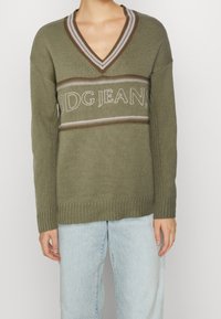 Olivově zelený pletený svetr s V-výstřihem, se kontrastním límcem v hnědé a bílé pruhované barvě a nápisem "BDGJEANS" přes hruď.