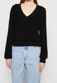 Schwarzer gerippter Pullover mit V-Ausschnitt, langen Ärmeln und cropped Passform, kombiniert mit hellblauen hoch taillierten Jeans.