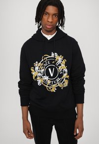 Juodas džemperis su gaubtu, turintis centrines puošnias baltas ir auksines detalės su užrašu "Versace Jeans Couture" ir stilizuota raide "V".