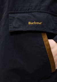 Κοντινό πλάνο σε τσέπη σκούρου μπουφάν με καφέ περιθώριο και κεντημένο το λογότυπο "Barbour" σε κίτρινο, με ένα χέρι που βρίσκεται μερικώς μέσα στην τσέπη.